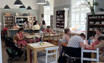 Group of people sitting at several tables in a bright pottery studio, painting ceramic mugs and dishes with brushes and colored paints; shelves with unpainted pottery and large windows are visible in the background.