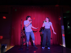 Two people stand on a small stage in front of a red curtain, each holding a microphone and facing each other while performing karaoke. Colored light spots are projected onto the curtain and floor. Two high stools and microphone stands are visible behind them.