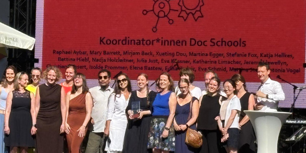 A group of approximately 15 people standing together on an outdoor stage. Behind them, a large red screen displays the text “Koordinator*innen Doc Schools” along with a list of names. One person in the center is holding an award. A podium and microphone stand are visible on the right side. The group is arranged in a line and facing the audience. The setting appears to be a formal event with stage lighting.